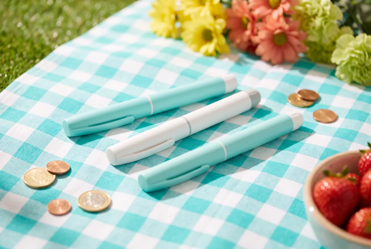 Plain injection pens in pastel colours with coins on a turquoise picnic blanket