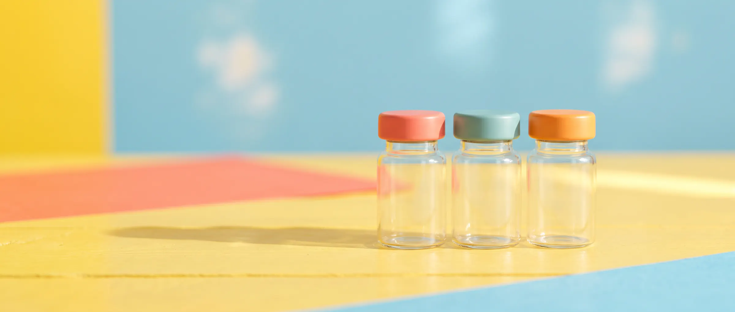 Three plain glass vials with coloured caps on a sunny yellow table